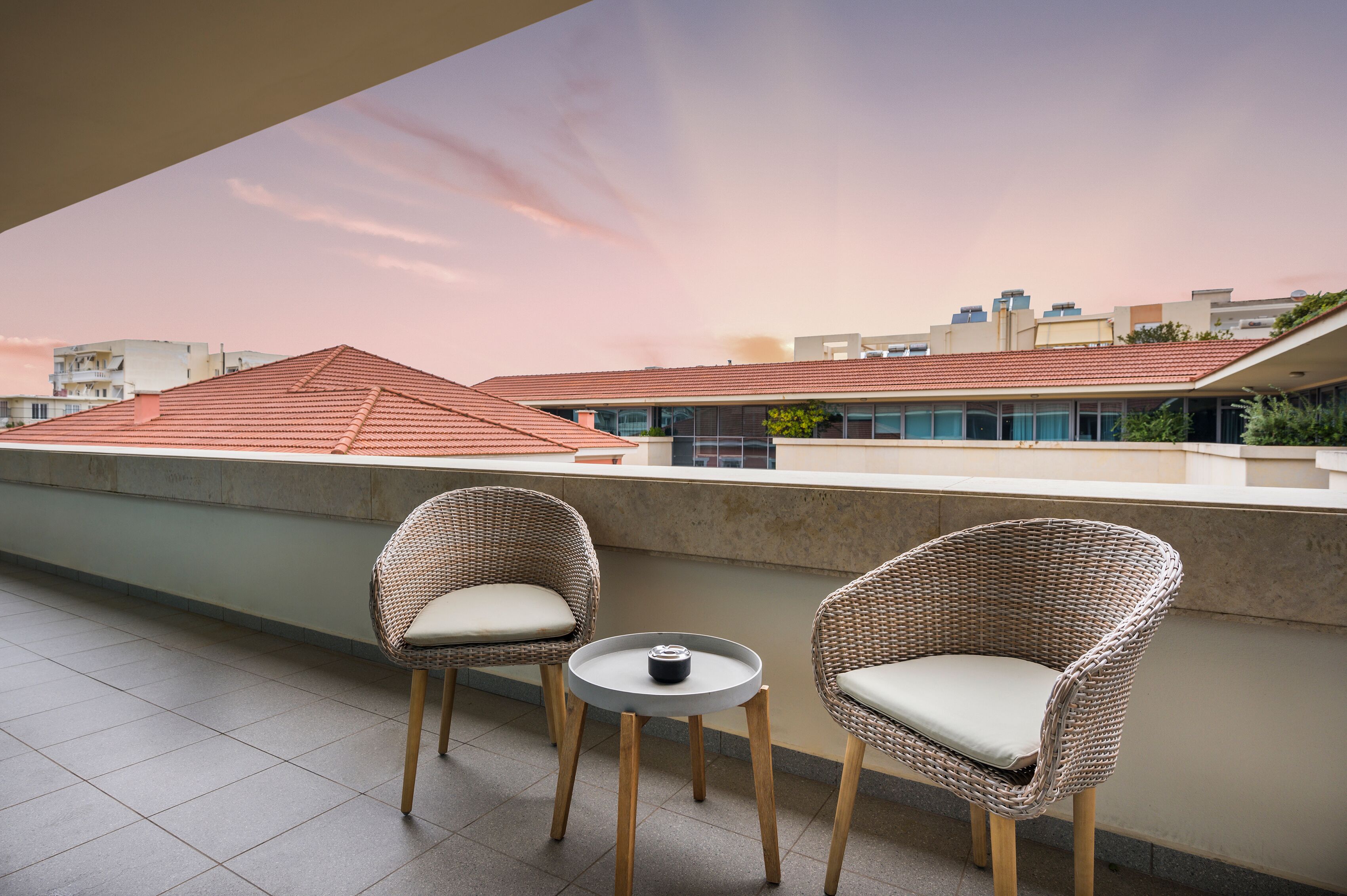 A balcony with two wicker chairs and a small round table, overlooking a cityscape with buildings and a sunset sky.