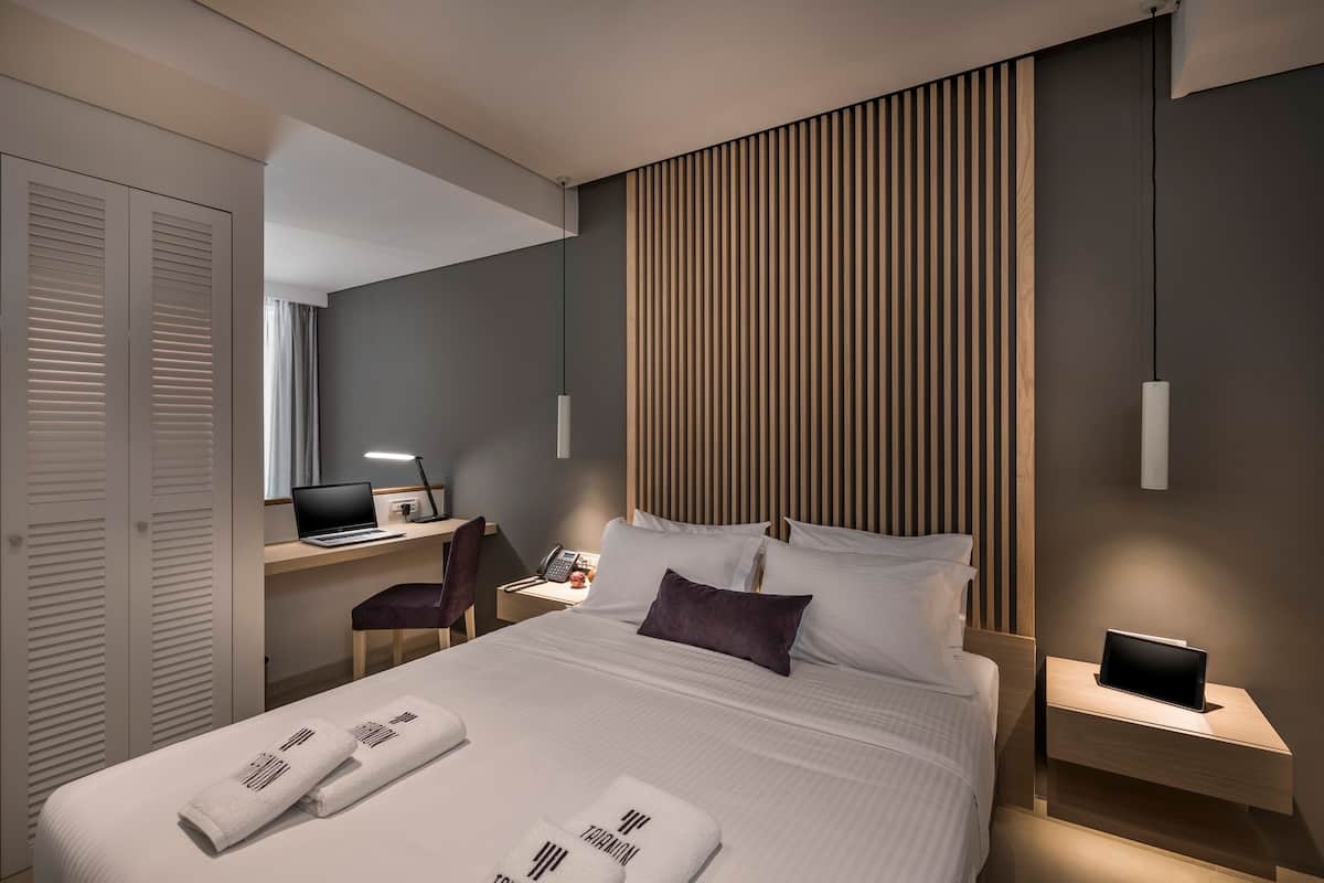 Una habitación de hotel moderna con una cama bien hecha, un escritorio con una computadora portátil, una silla y una mesita de noche con una tableta.