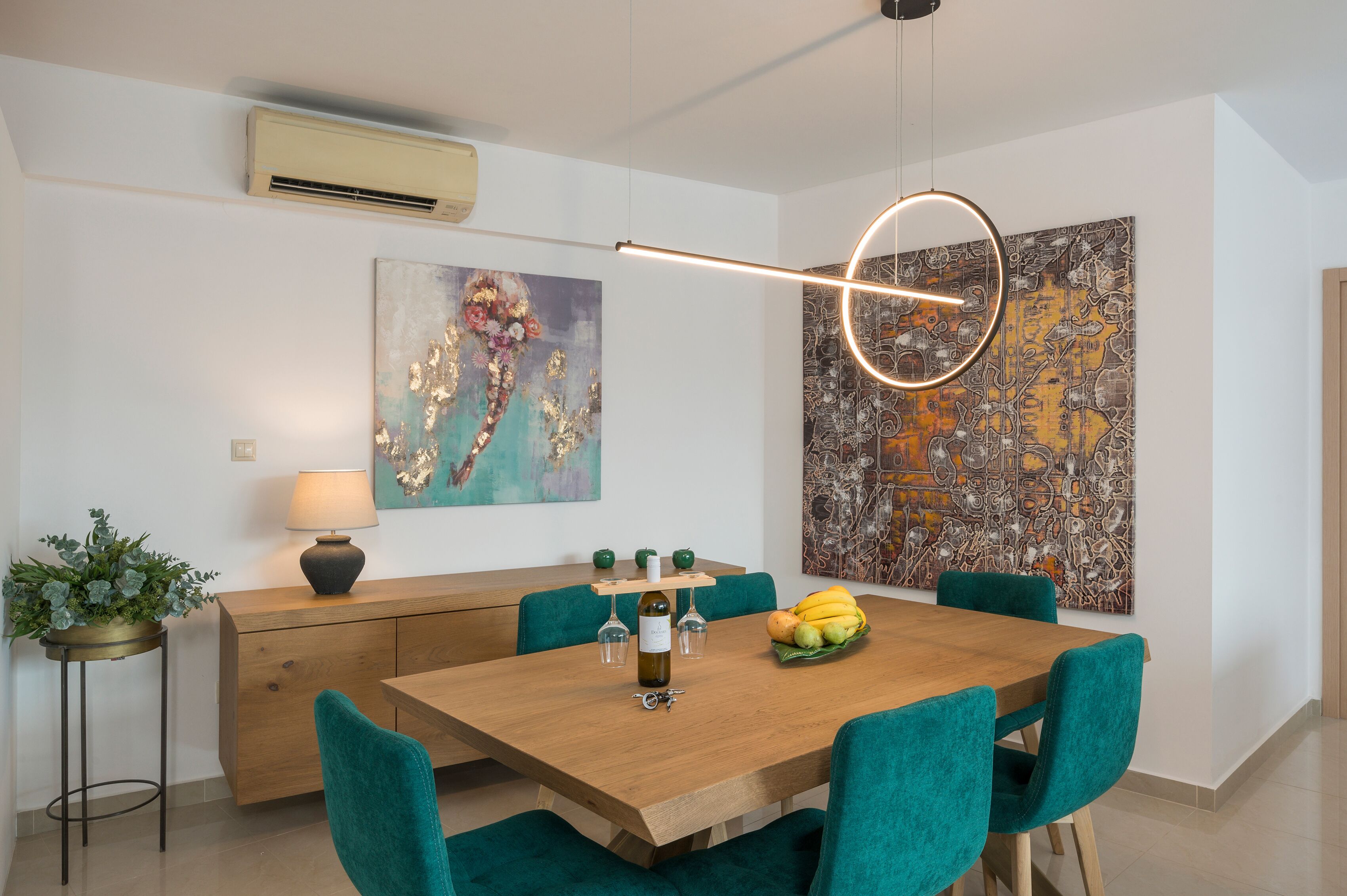 Uma sala de jantar com uma mesa de madeira, cadeiras cor turquesa, uma planta em vaso, uma garrafa de vinho e uma cesta de frutas.