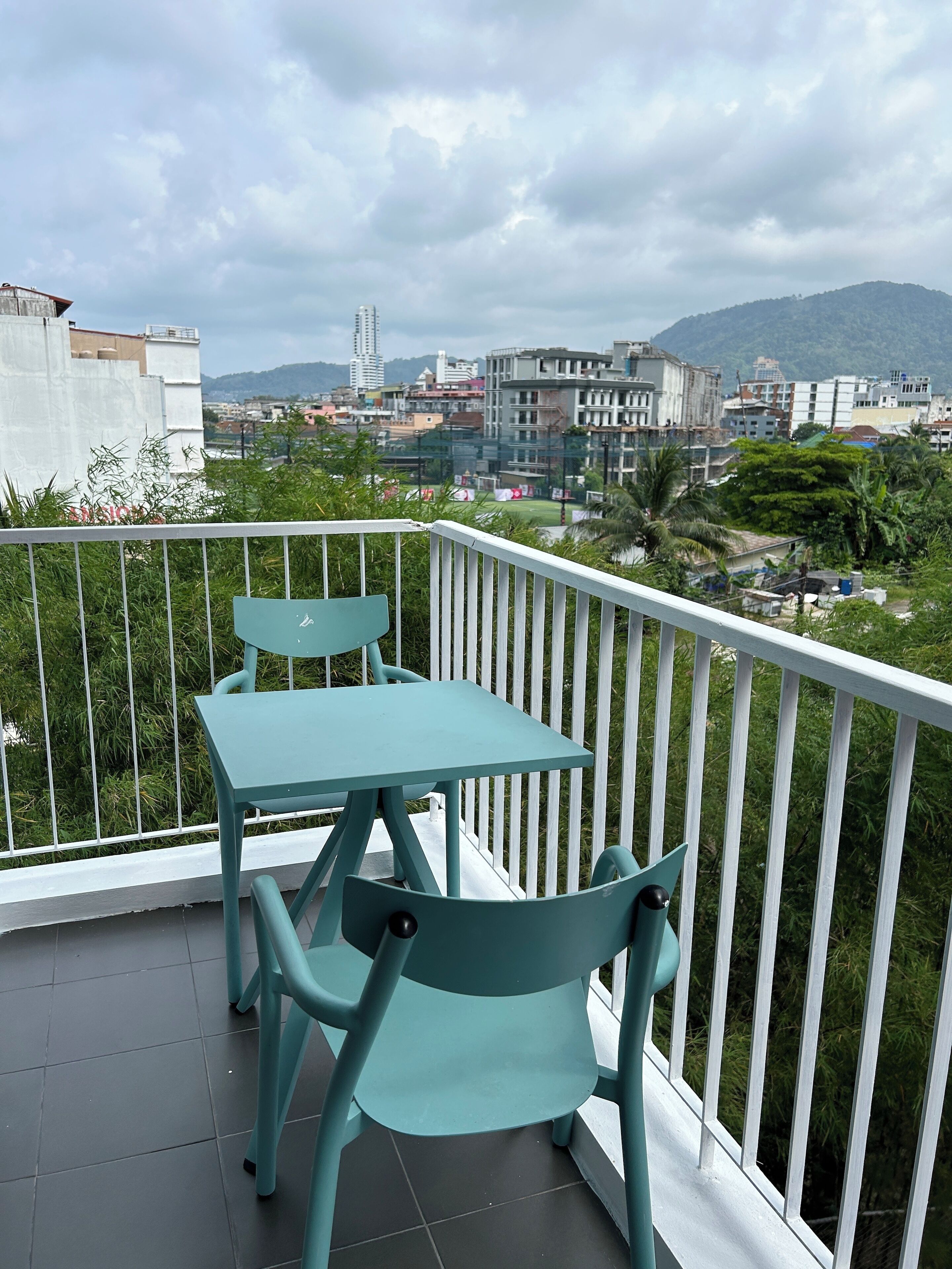 Varanda com duas cadeiras turquesa e uma mesa, com vista para um horizonte urbano com edifícios e montanhas.