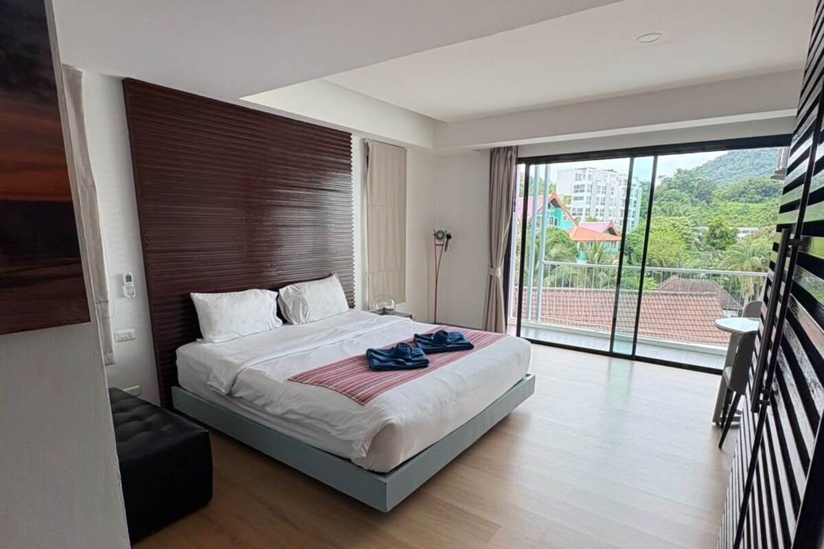 A modern bedroom with a large bed, a nightstand, a standing lamp, and a view of buildings through the window.
