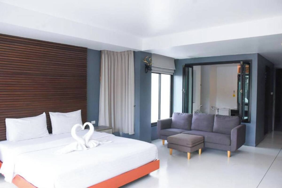 A modern hotel room with a large bed, a grey sofa, and a small ottoman.