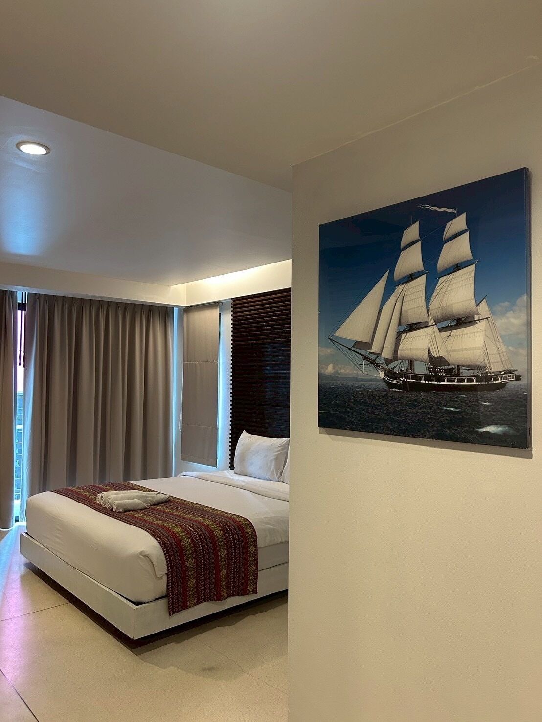 Ein modernes Hotelzimmer mit einem großen Bett, einem Fenster mit Vorhängen und einem Gemälde eines Segelschiffs an der Wand.