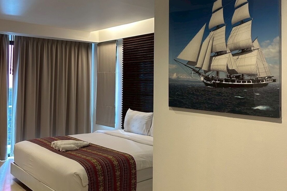 A modern hotel room with a large bed, a window with curtains, and a painting of a sailing ship on the wall.