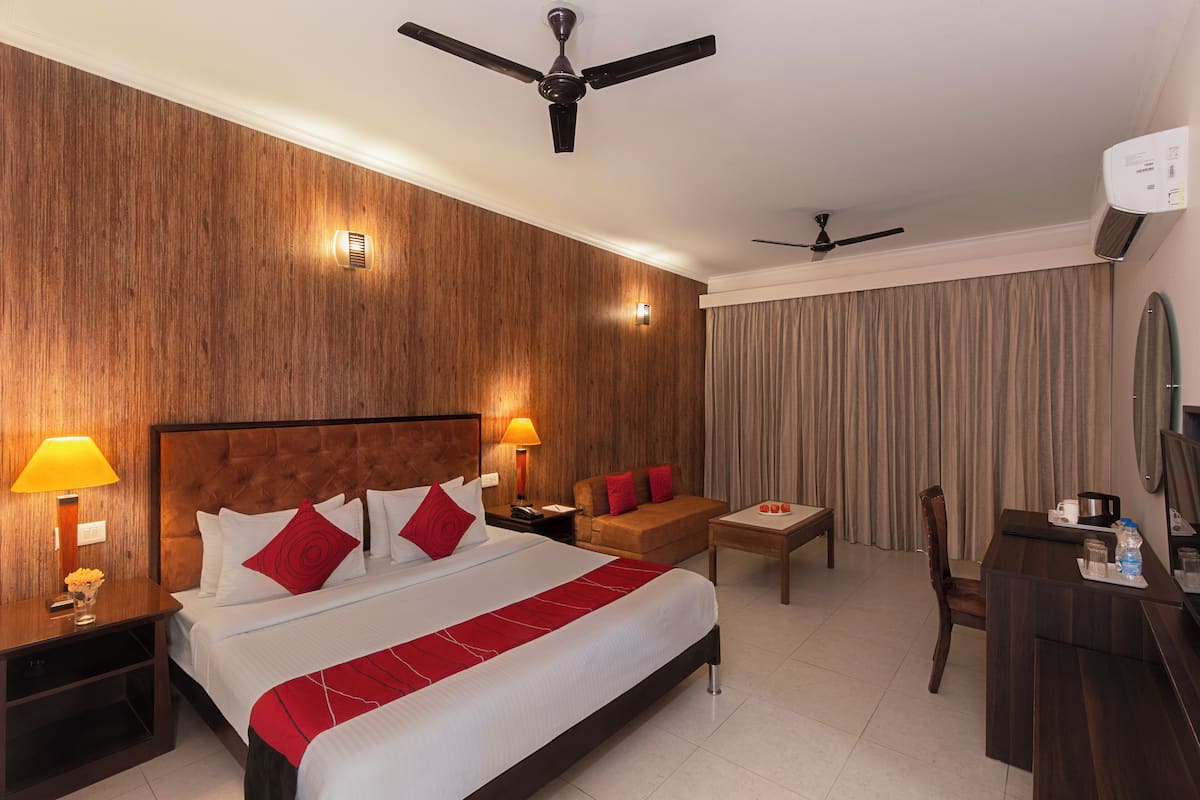 A hotel room with a large bed, two bedside tables with lamps, a desk, a chair, a ceiling fan, and a window with curtains.