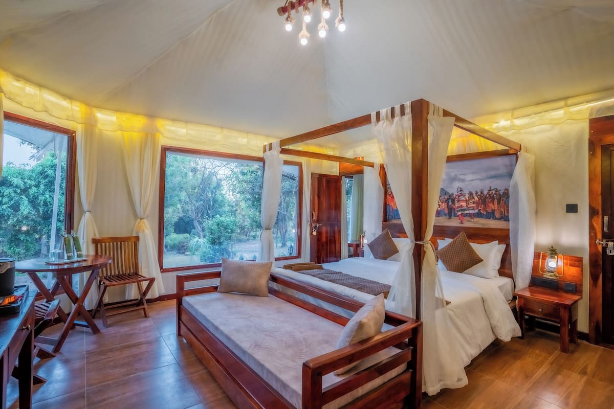 A four-poster bed with a canopy, a wooden desk, and a chair in a room with large windows and a view of greenery.
