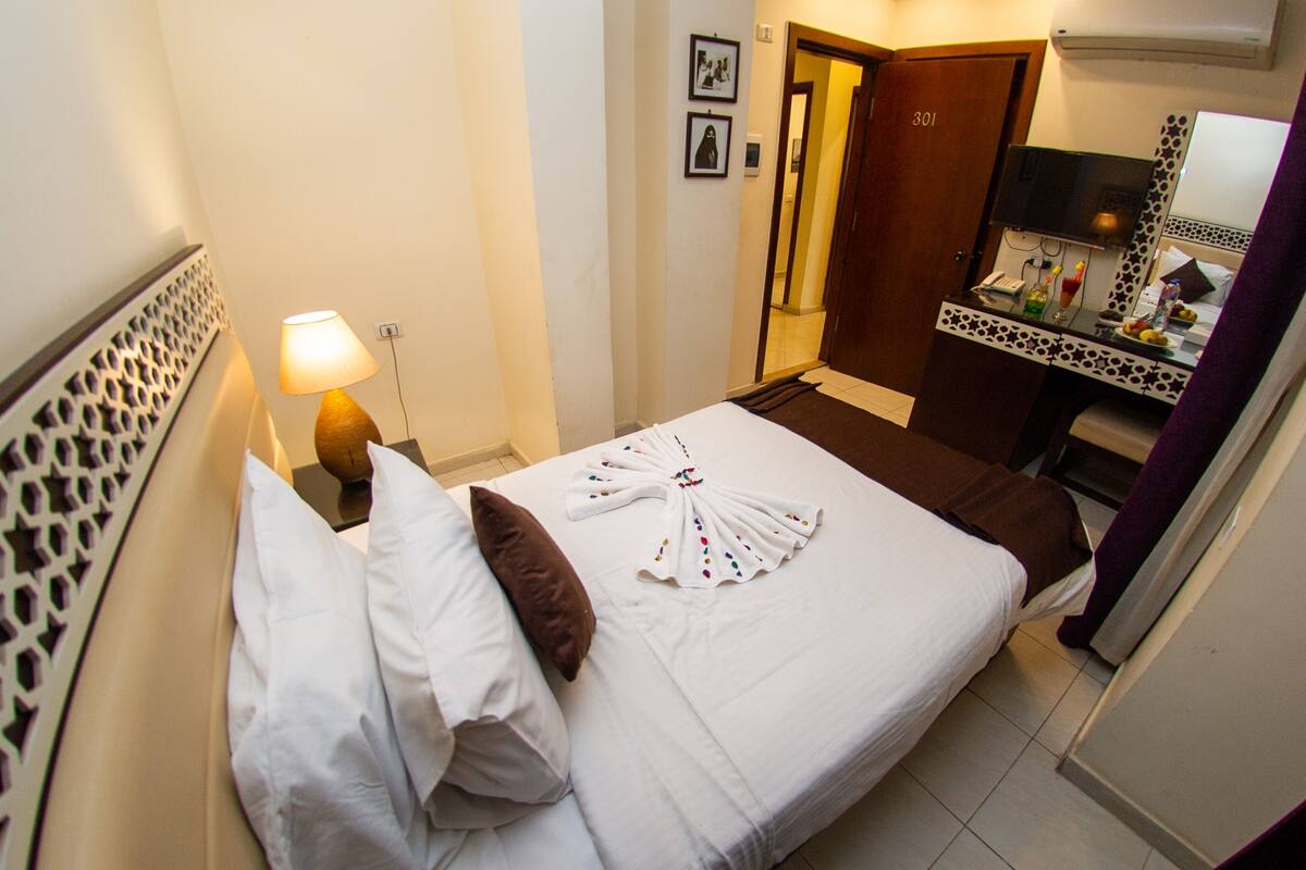 A hotel room with a neatly made bed, a bedside lamp, a small table with toiletries, and a mirror with a decorative frame.