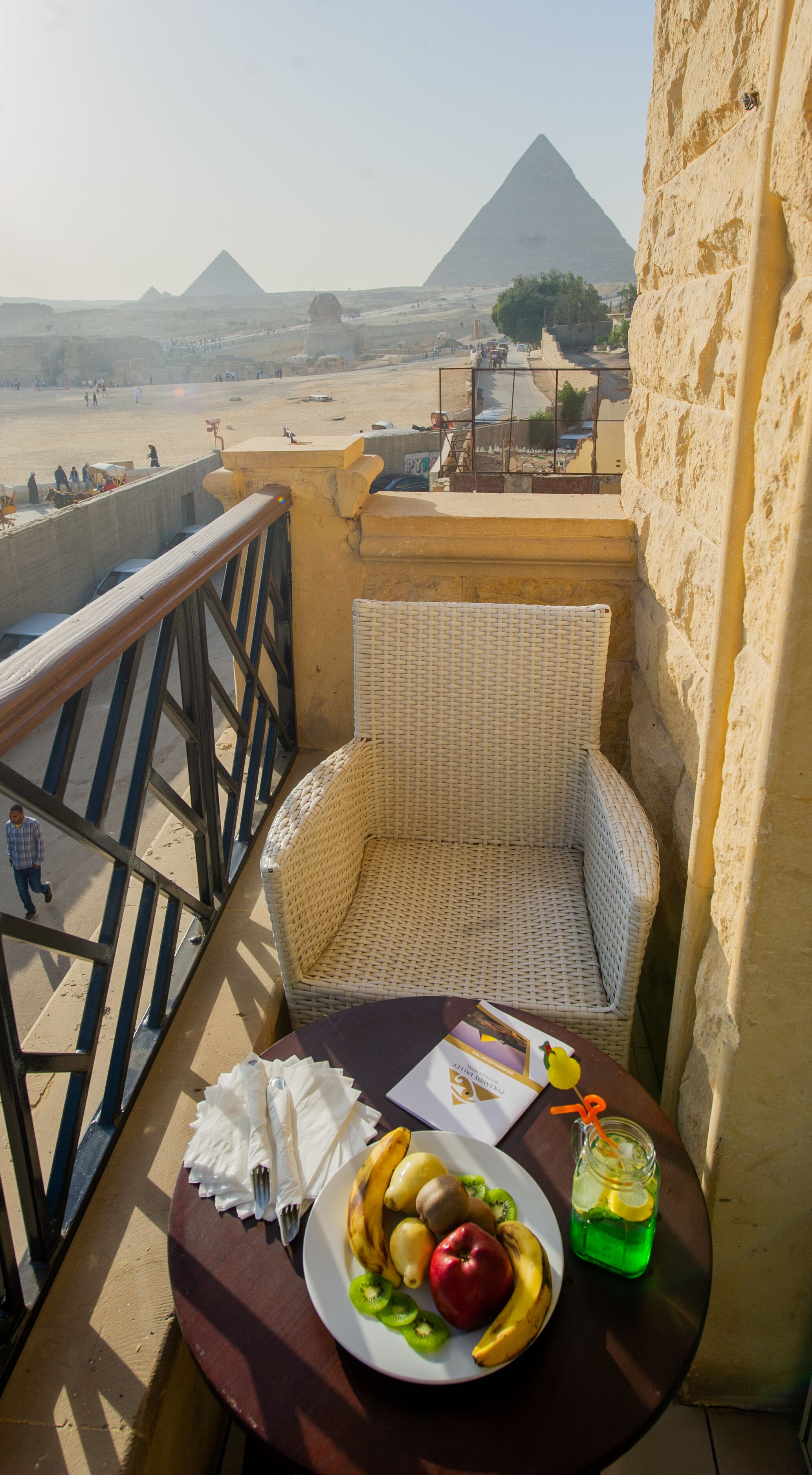 A balcony with a wicker chair, a table with a plate of fruit, a drink, and a brochure. A pyramid is visible in the background.