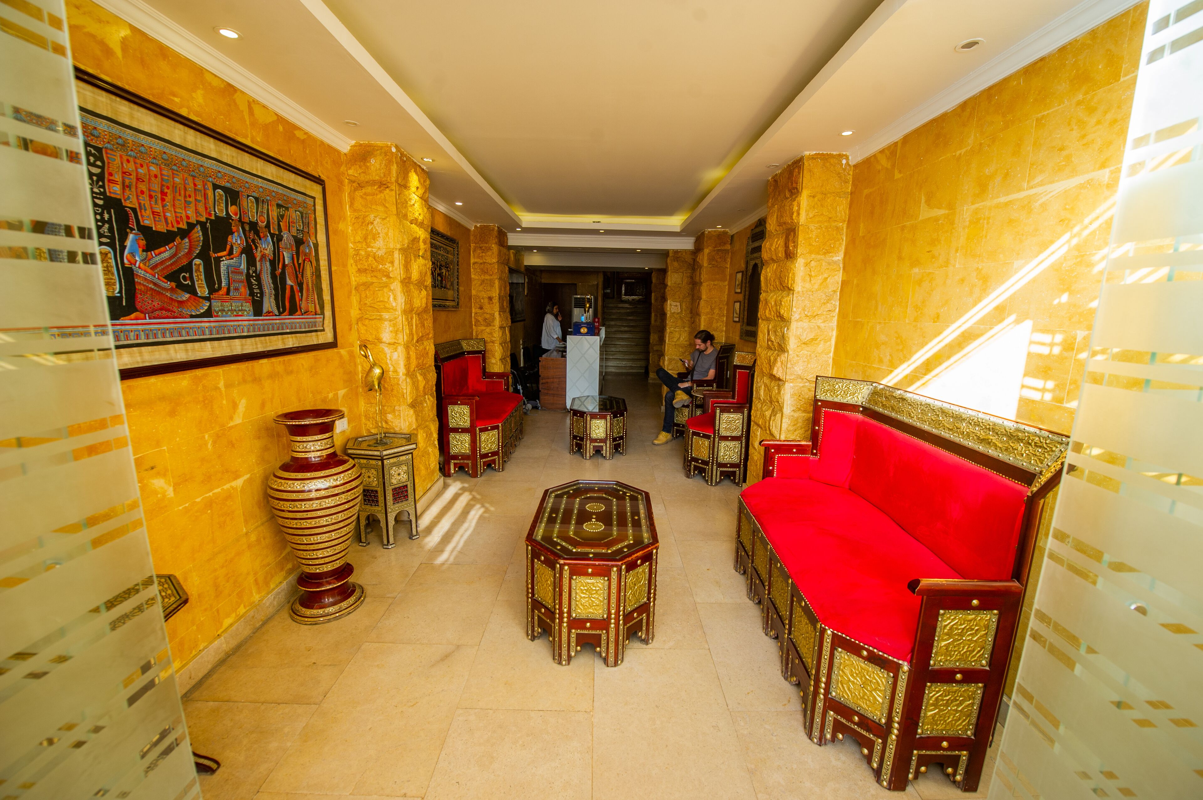 Un vestíbulo con un sofá rojo, una mesa de centro y un jarrón decorativo. Hay un cuadro en la pared y una escalera al fondo.