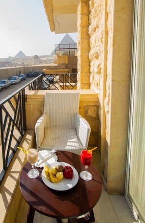 A balcony with a wicker chair, a table with a plate of fruit and two drinks, and a view of the pyramids in the background.
