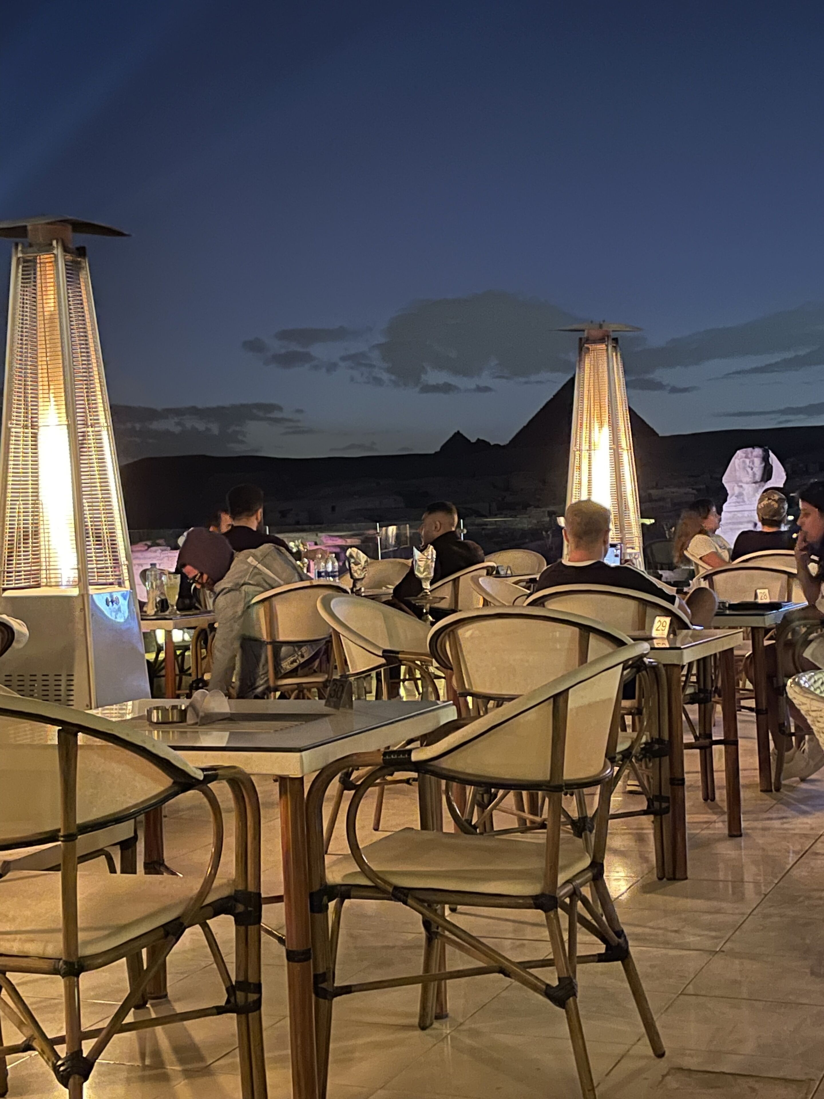 An outdoor dining area with tables and chairs, a view of the mountains, and a large illuminated structure in the background.