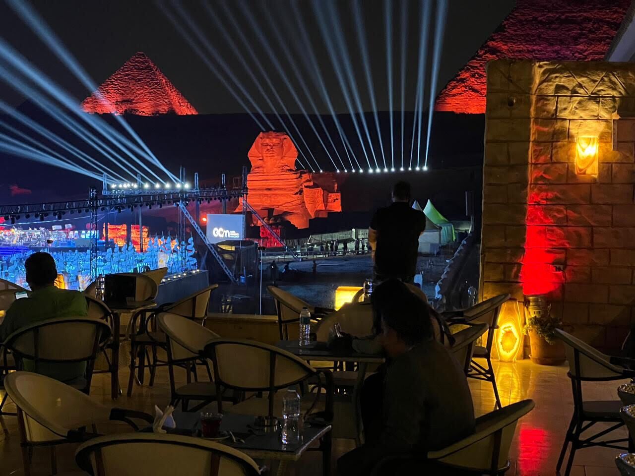 An outdoor dining area with tables and chairs, a stage with a performer, and a large illuminated structure in the background.