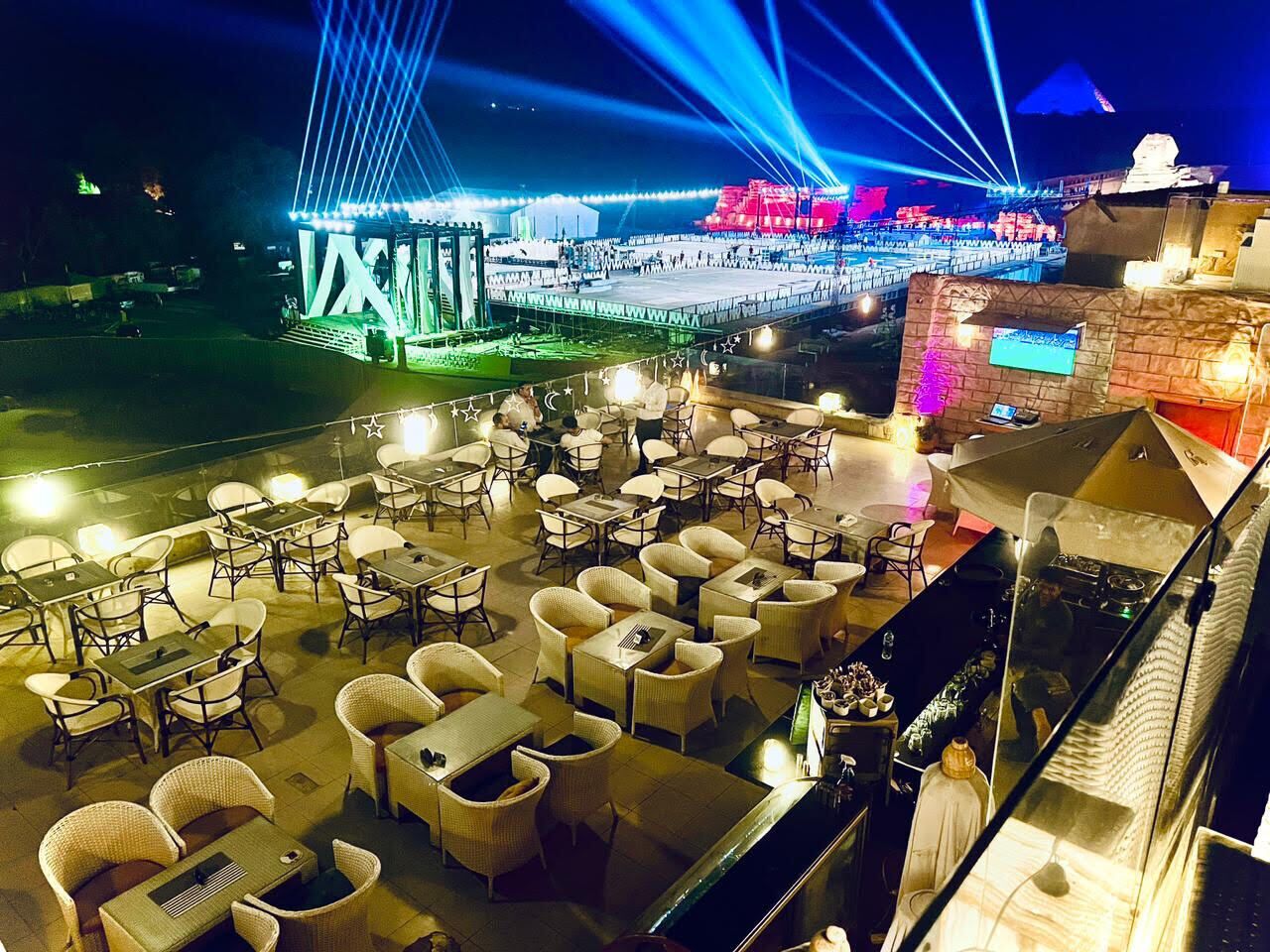 An outdoor dining area with wicker furniture and a view of a brightly lit stage and bridge.