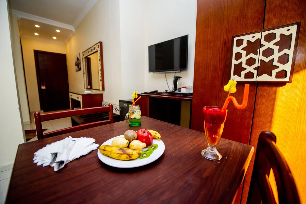 A dining table with a plate of fruit, a glass of juice, and a napkin. A television is mounted on the wall, and there is a wooden cabinet with a decorative panel.