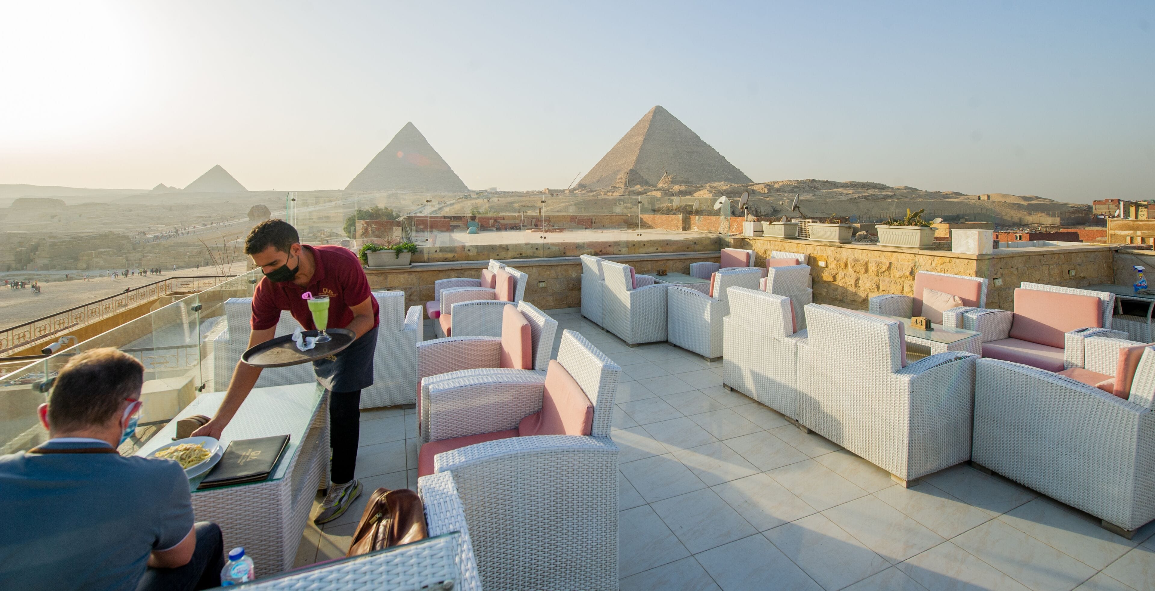 A rooftop terrace with wicker furniture, a waiter serving a guest, and a view of the pyramids in the background.