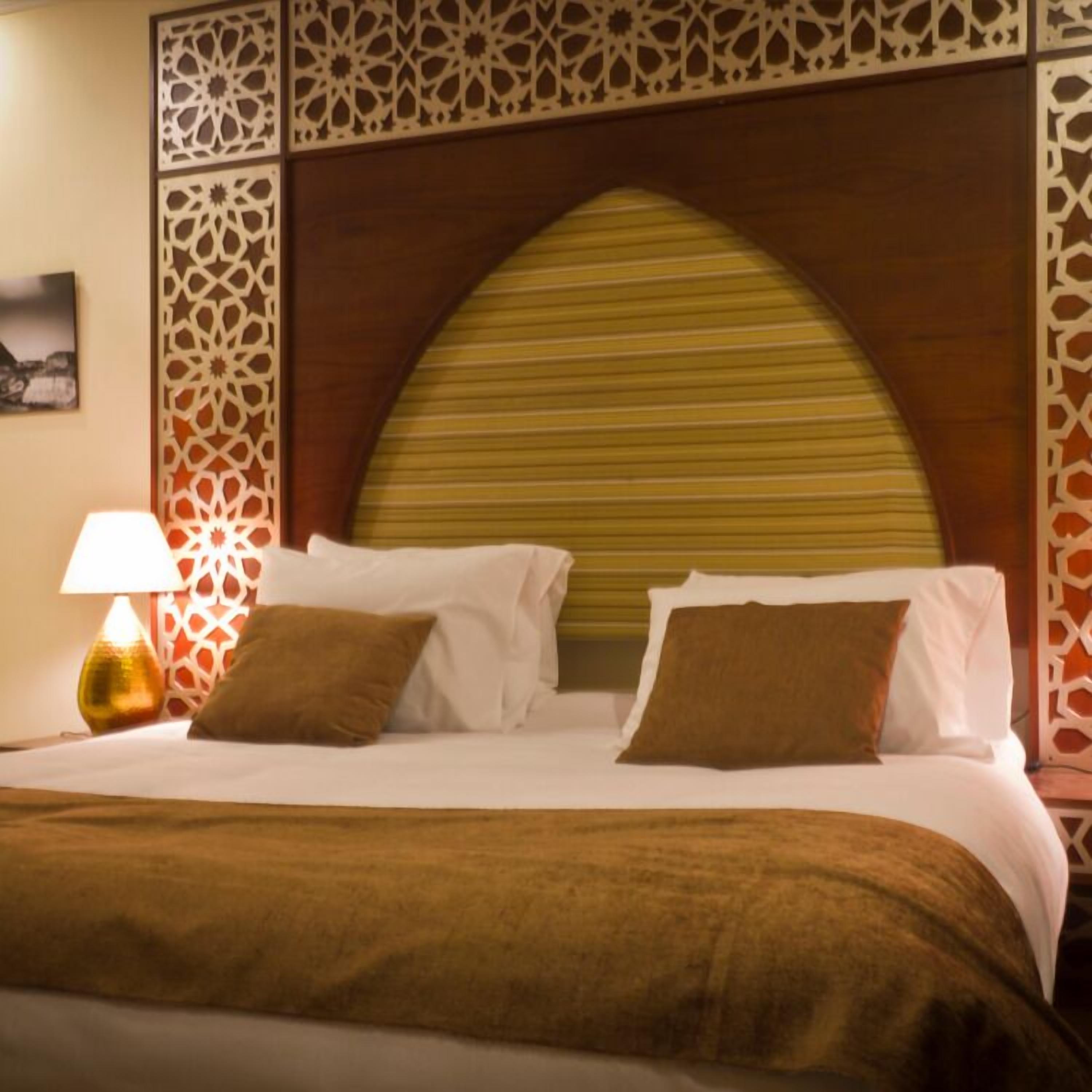 Uma cama bem arrumada com lençóis brancos e almofadas marrons, um painel decorativo na parede com padrões intrincados e um abajur com base dourada.