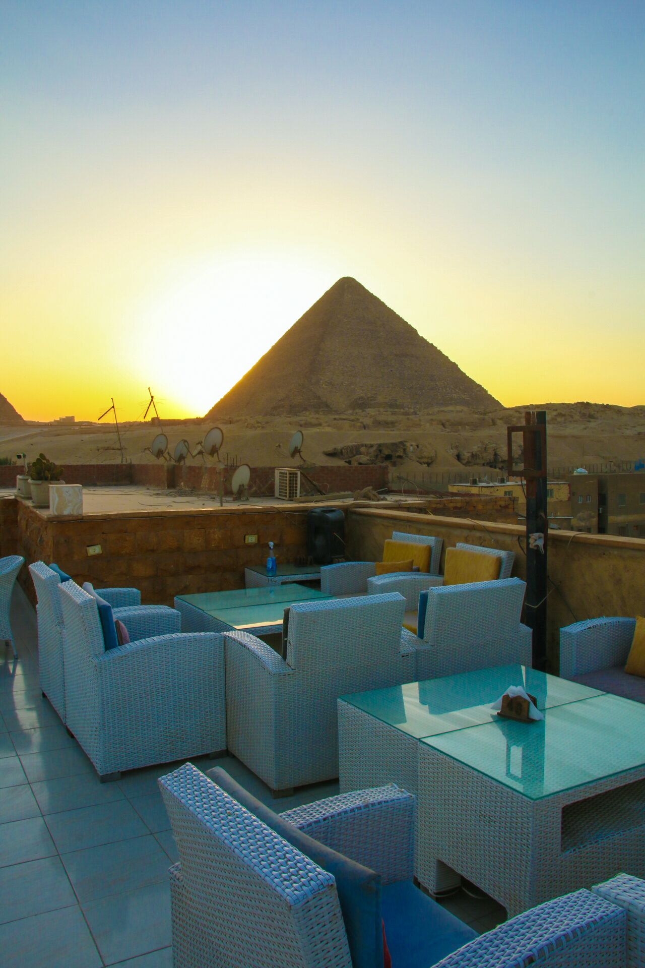 Une terrasse sur le toit, aménagée avec des meubles en osier et une table en verre, offrant une vue sur une pyramide et un paysage désertique.