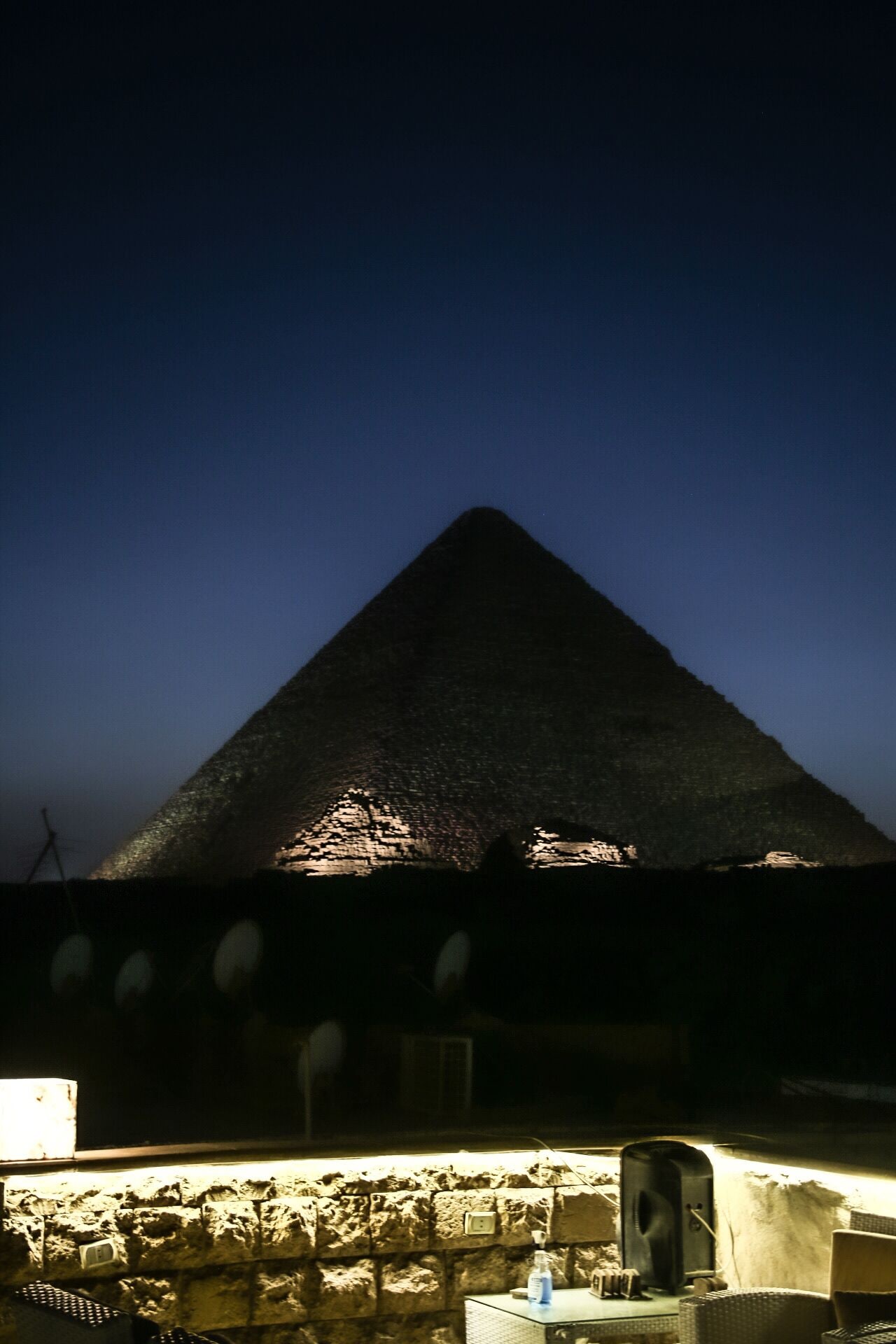 A pyramid-shaped structure illuminated at night, with a stone wall and a small table with a bottle in the foreground.