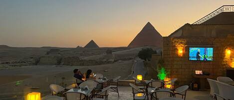 An outdoor dining area with tables and chairs, a TV screen, and a view of the pyramids.