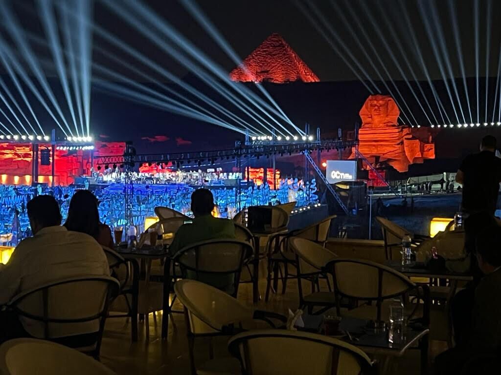 A restaurant with tables and chairs, a stage with a large screen, and a mountain-like structure in the background.