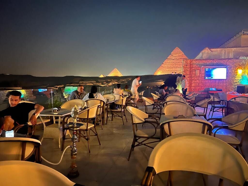 Un área de comedor al aire libre con mesas y sillas, un narguilé y vistas a las pirámides al fondo.