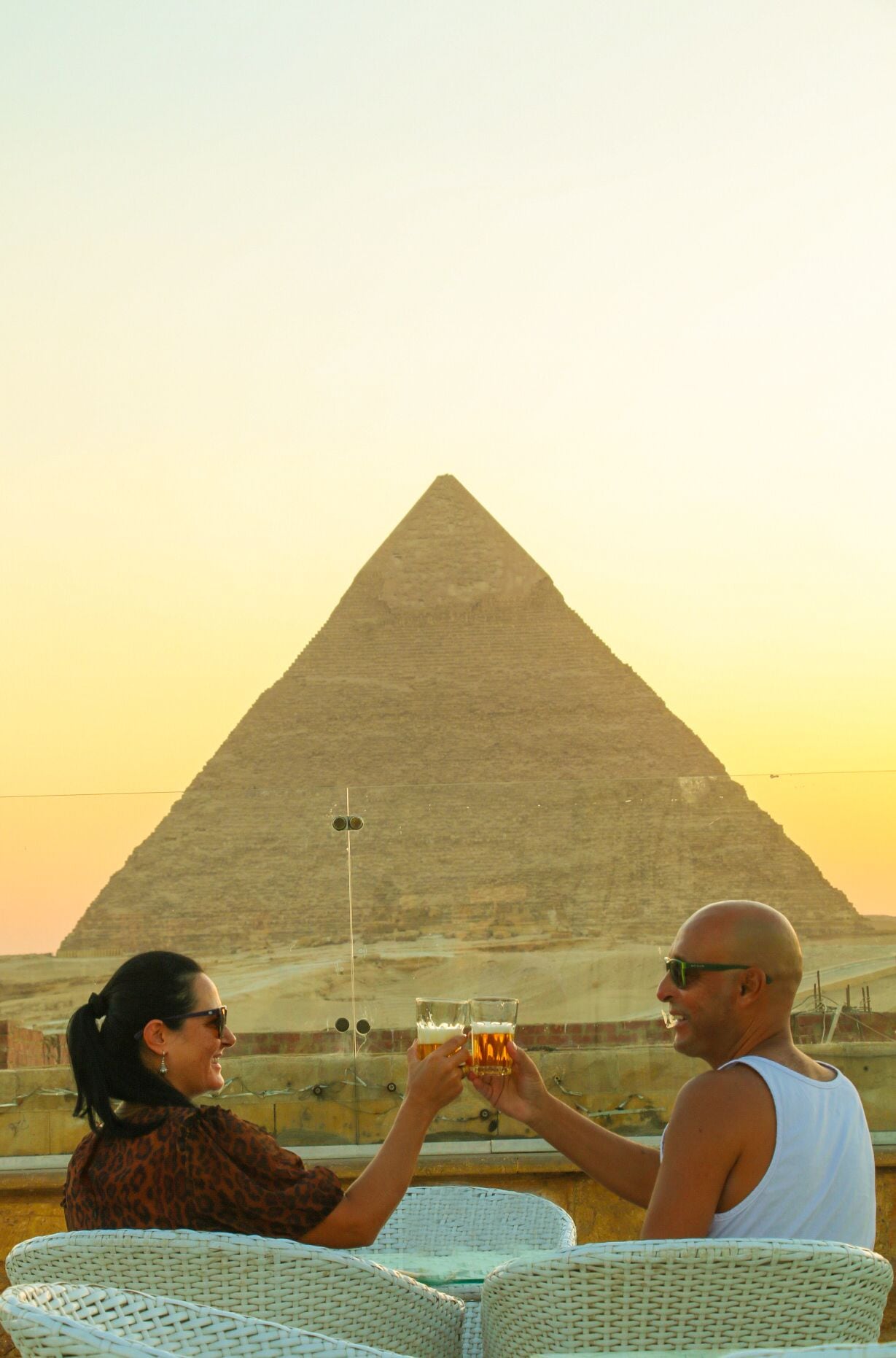 Two people toasting with drinks in front of the Great Pyramid of Giza.