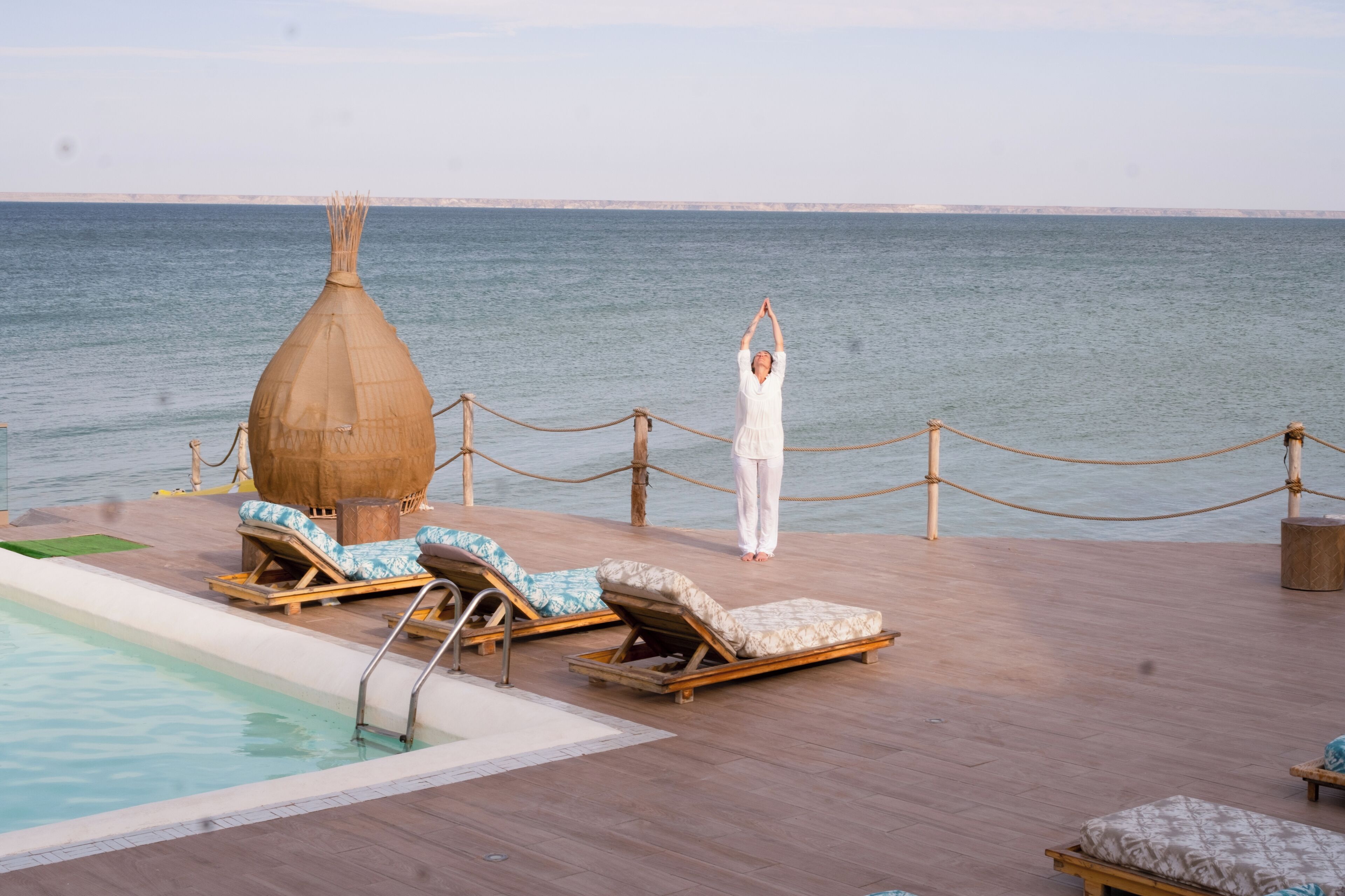 Zona de piscina con sillones de descanso y una estructura de madera, con vistas al mar tranquilo.
