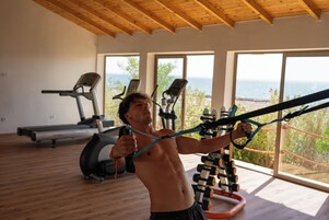 Un gimnasio con equipo de ejercicio, amplias ventanas y vista al mar.