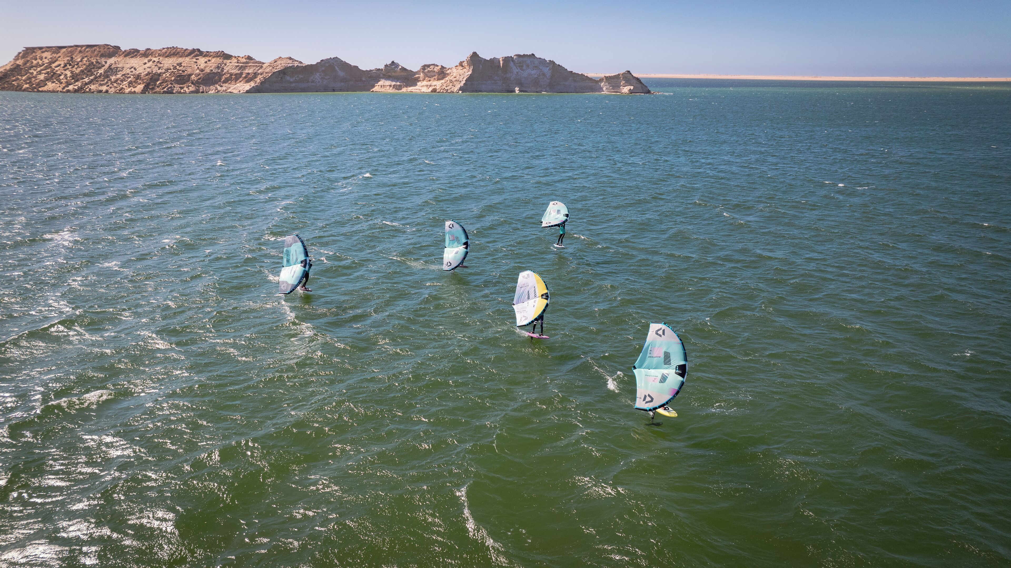 A group of windsurfers on a calm sea with rocky cliffs in the background.