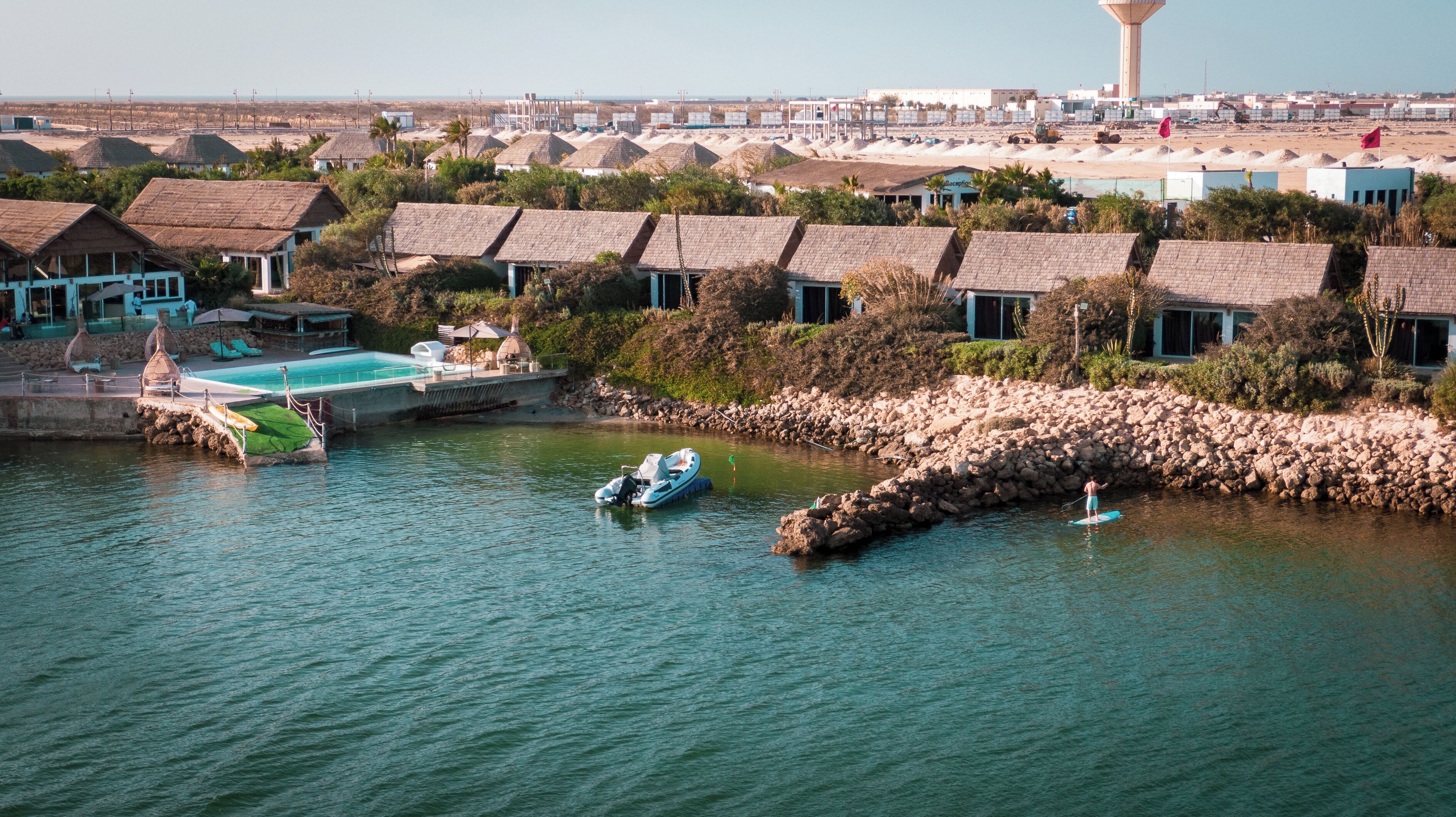Un complejo turístico frente al mar con bungalows sobre el agua, piscina y un litoral rocoso.