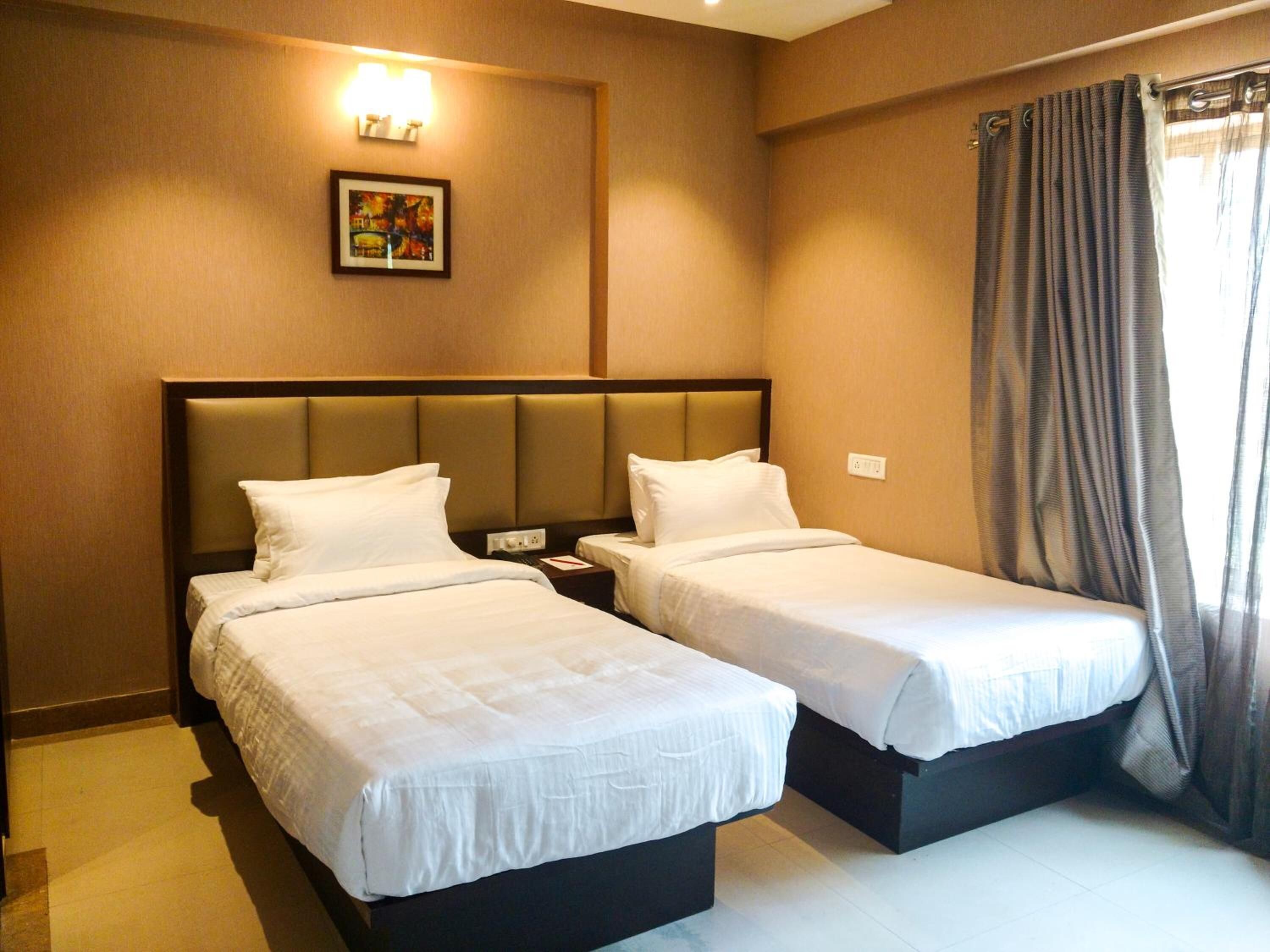 A hotel room with two single beds, a headboard, a nightstand, a wall-mounted light, and a window with curtains.