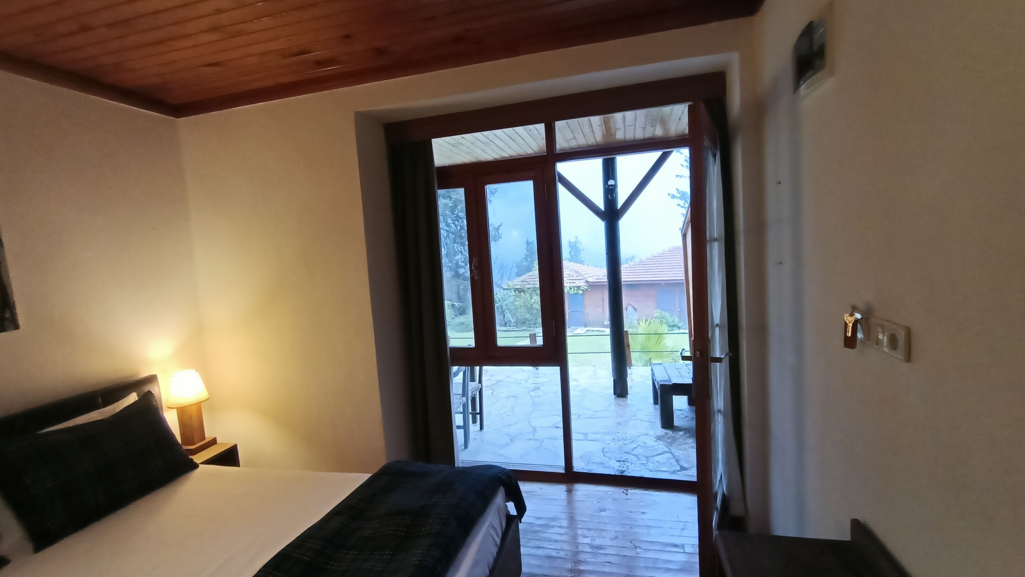 Un dormitorio con cama, mesita de noche con lámpara y puerta corrediza de vidrio que da a un patio exterior con mesa y sillas.