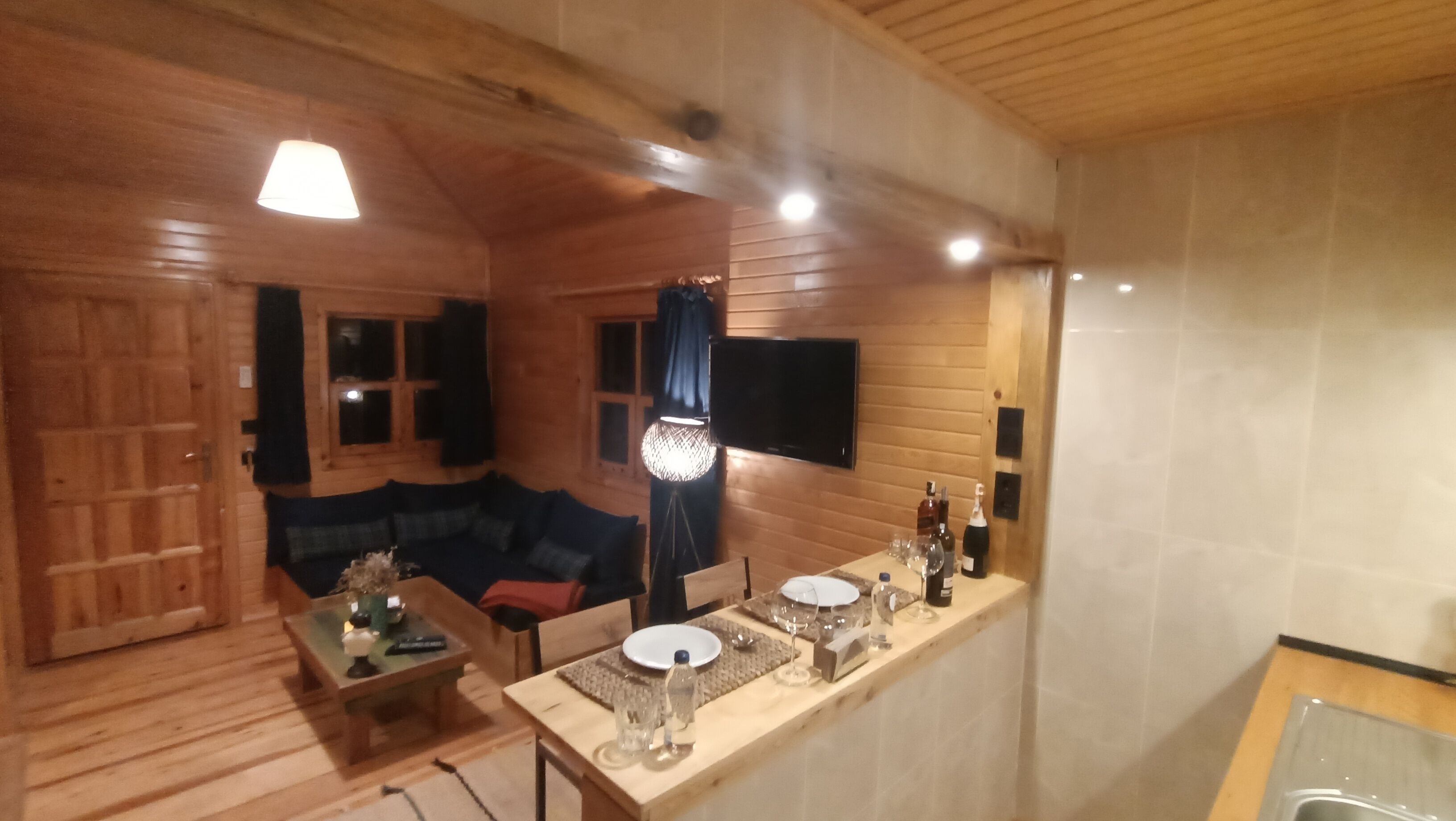 Interior acogedor de una cabaña con suelo de madera, un sofá, una mesa de comedor con platos y vasos, y un televisor montado en la pared.
