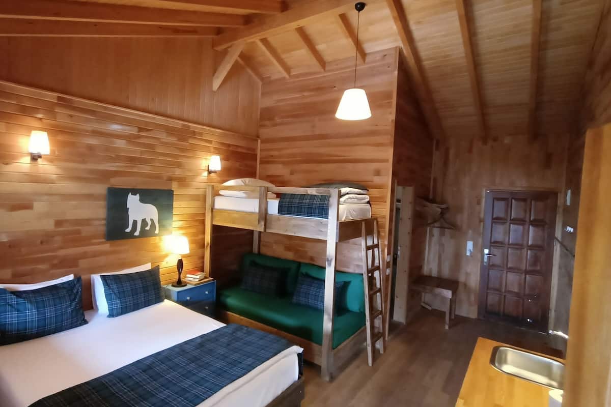 Una habitación de cabaña de madera con literas, una cama con almohadas, una lámpara de pared y techo de madera.