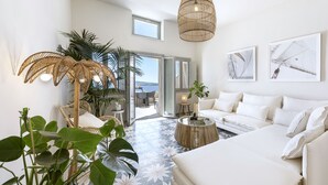 A spacious living room with a large white sofa, a round coffee table, and a wicker lamp. There are several potted plants and a view of the outdoors through a glass door. - Caldera Houses (Santorini)