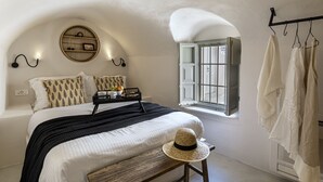 A bedroom with a bed, a round wall shelf, a window with shutters, and a coat rack. - Caldera Houses (Santorini)