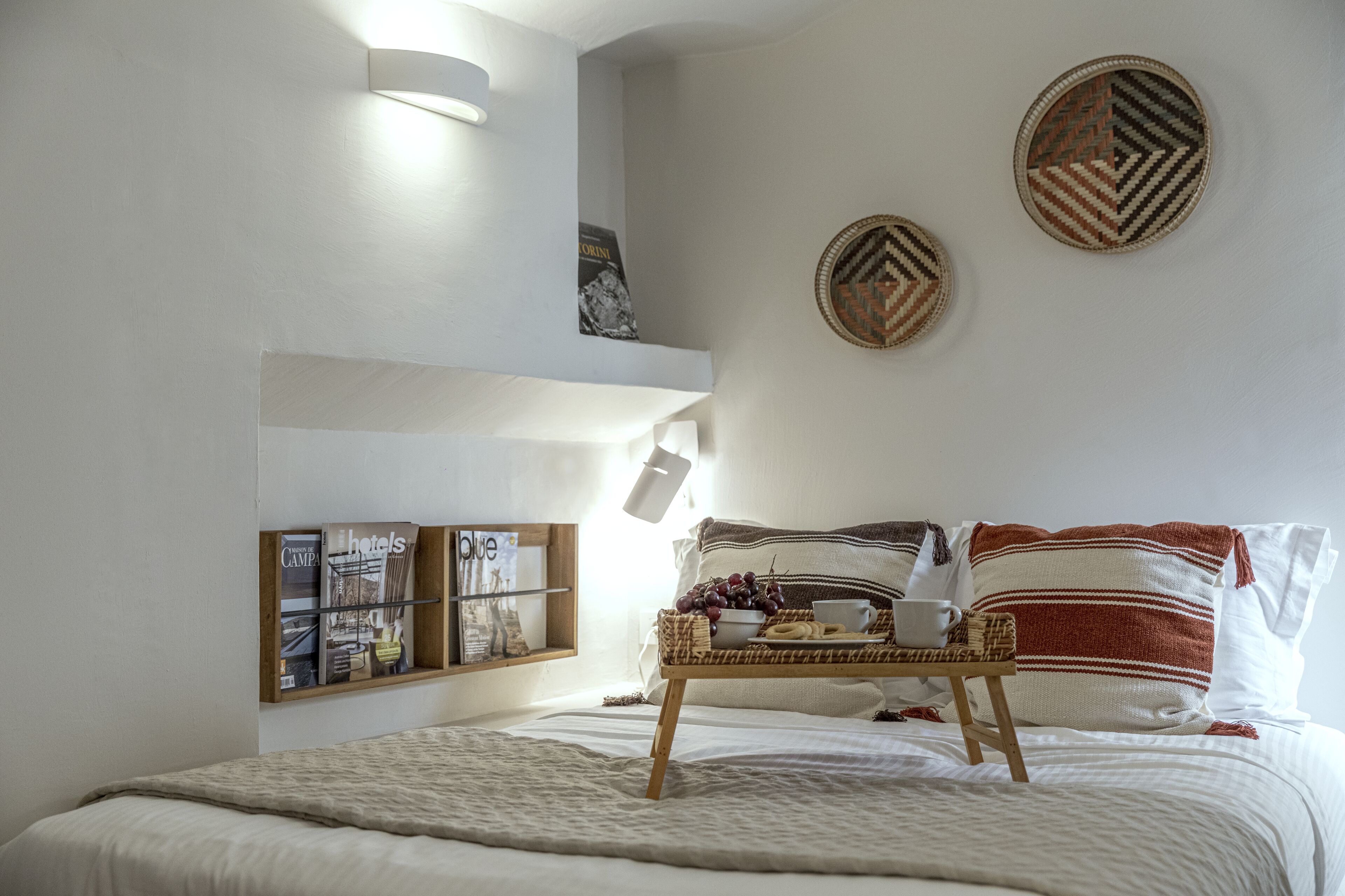 A neatly arranged bedroom with a bed, a bedside table with a tray of fruit and a cup, and a wall-mounted shelf with magazines.