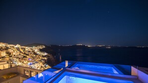 A rooftop with a glass-enclosed hot tub overlooking a coastal town at night. - Caldera Houses (Santorini)