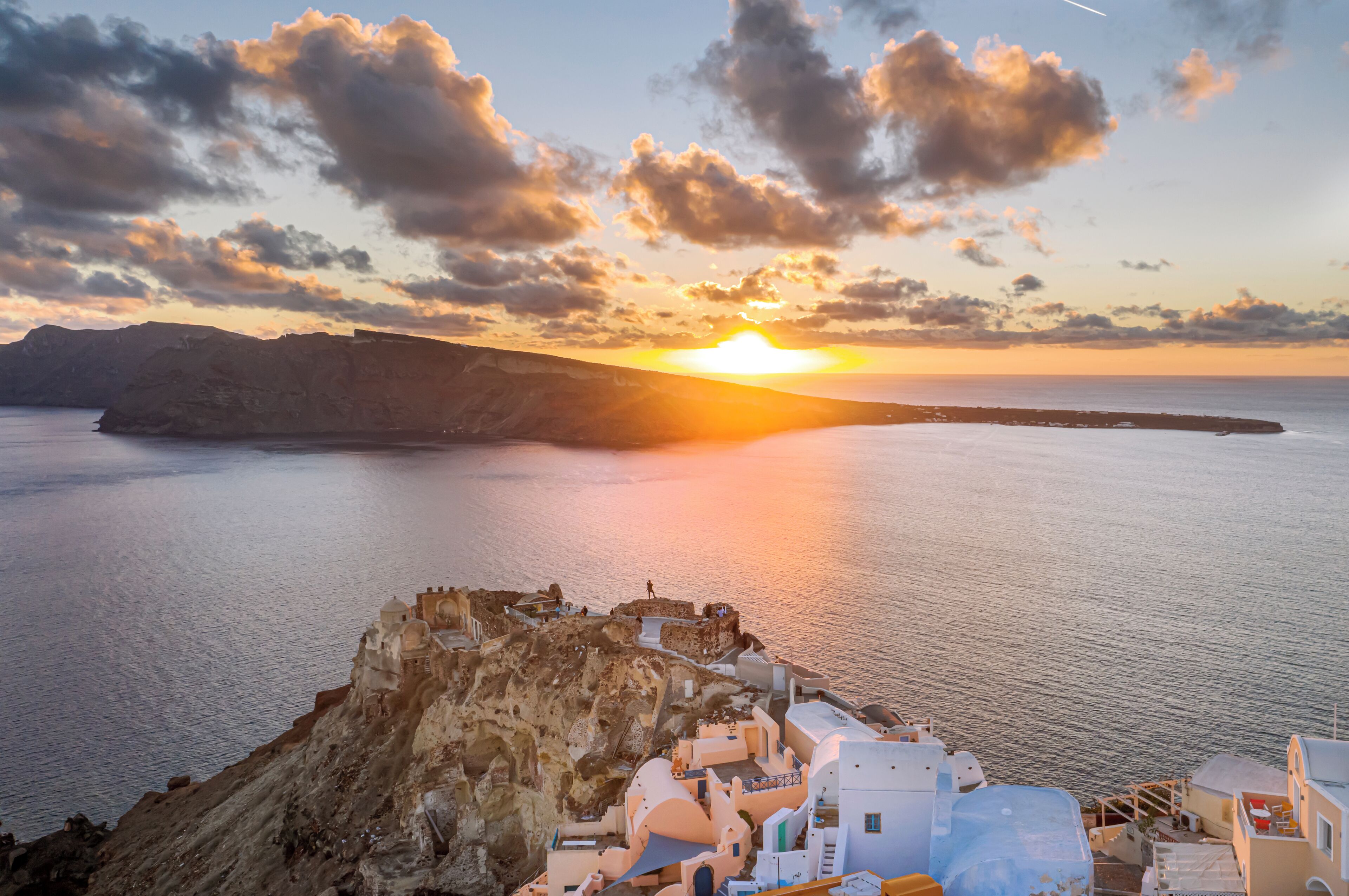 A coastal village with white buildings and a cliff overlooking the sea at sunset.