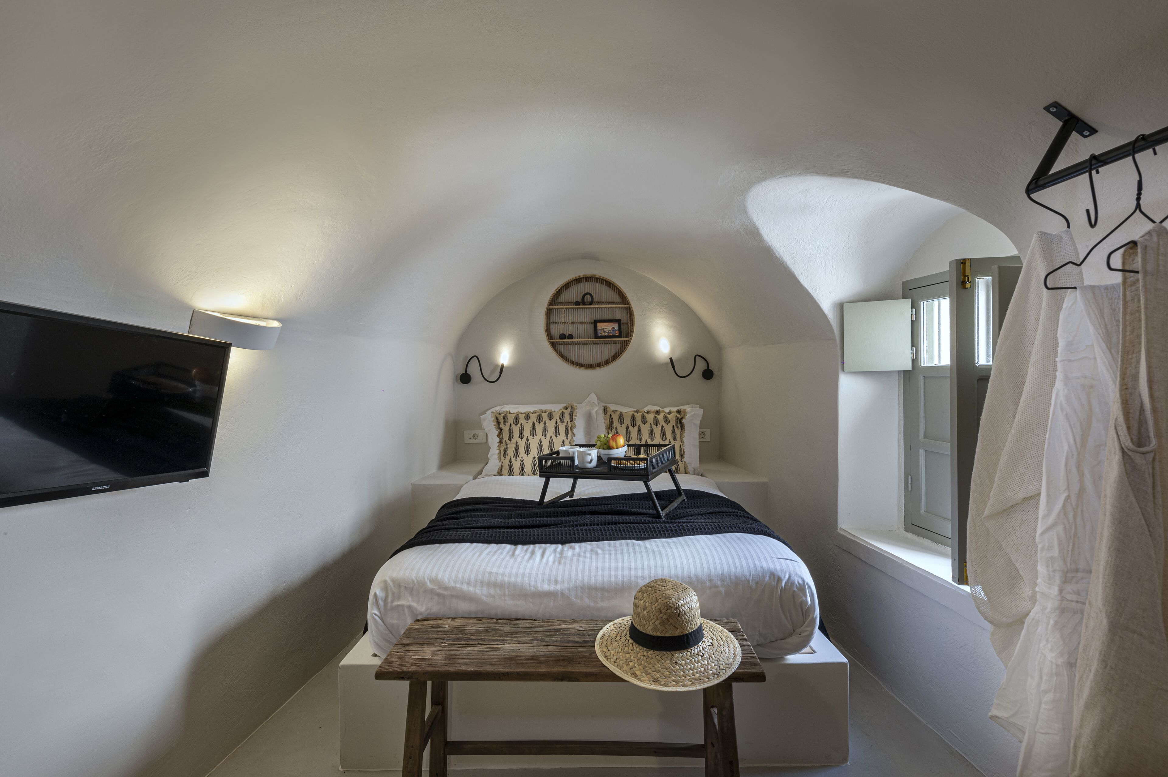 A bedroom with a bed, a bedside table, a hat, a wall-mounted TV, and a window with curtains.