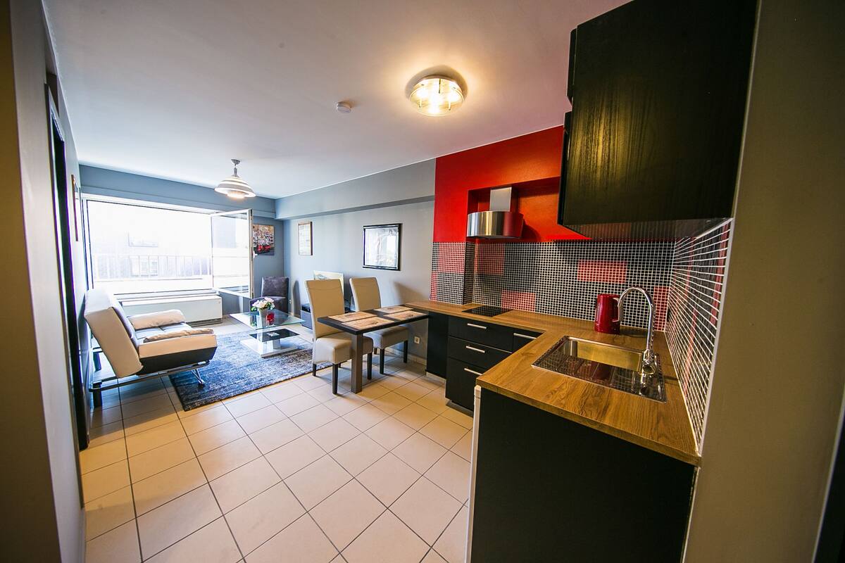 A modern kitchen with a wooden countertop, a sink, and a red backsplash. There is a dining area with a table and chairs, and a sofa in the background.