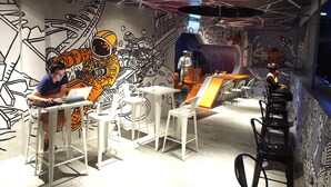 A room with a mural of astronauts and space elements, a white table with a laptop, and a VR headset on a table. - Space Hotel (Kuala Lumpur)