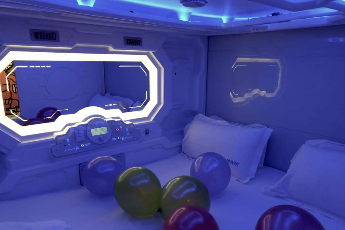 A room with a futuristic design, featuring a control panel, a bed with white bedding, and several balloons.