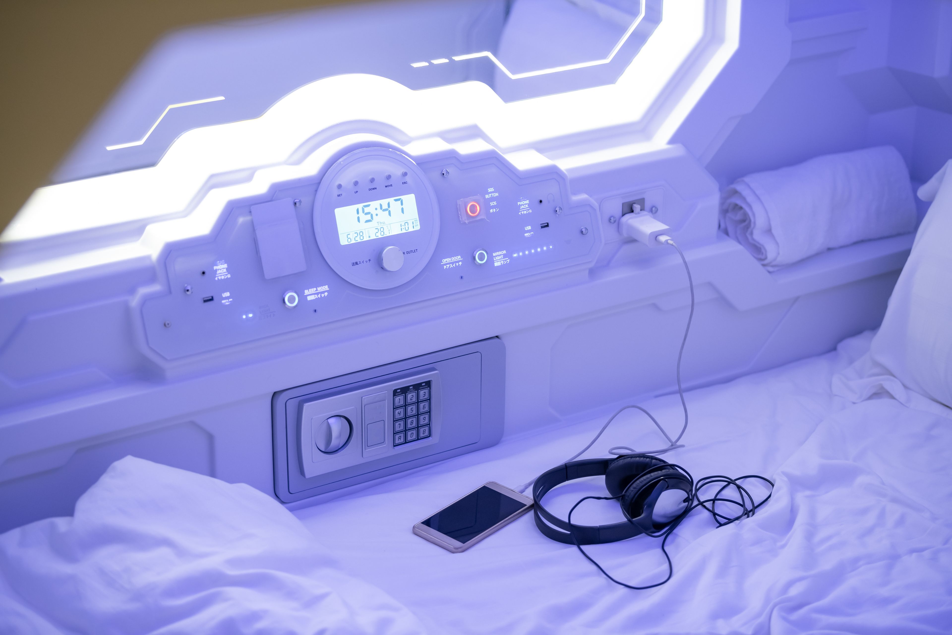 A bed with a digital clock, a control panel, a smartphone, and headphones.