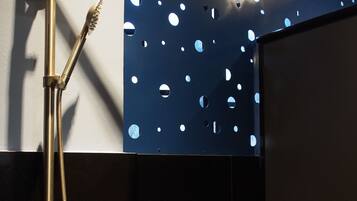 A modern shower with a handheld showerhead and a fixed rain showerhead, a handheld toothbrush, and a soap dispenser on a black tiled wall.