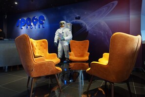 Un espace d’accueil avec un décor sur le thème de l’espace, comprenant une grande statue d’astronaute et une enseigne sur laquelle on peut lire « Hôtel spatial ». Deux chaises orange sont disposées autour d’une petite table ronde.