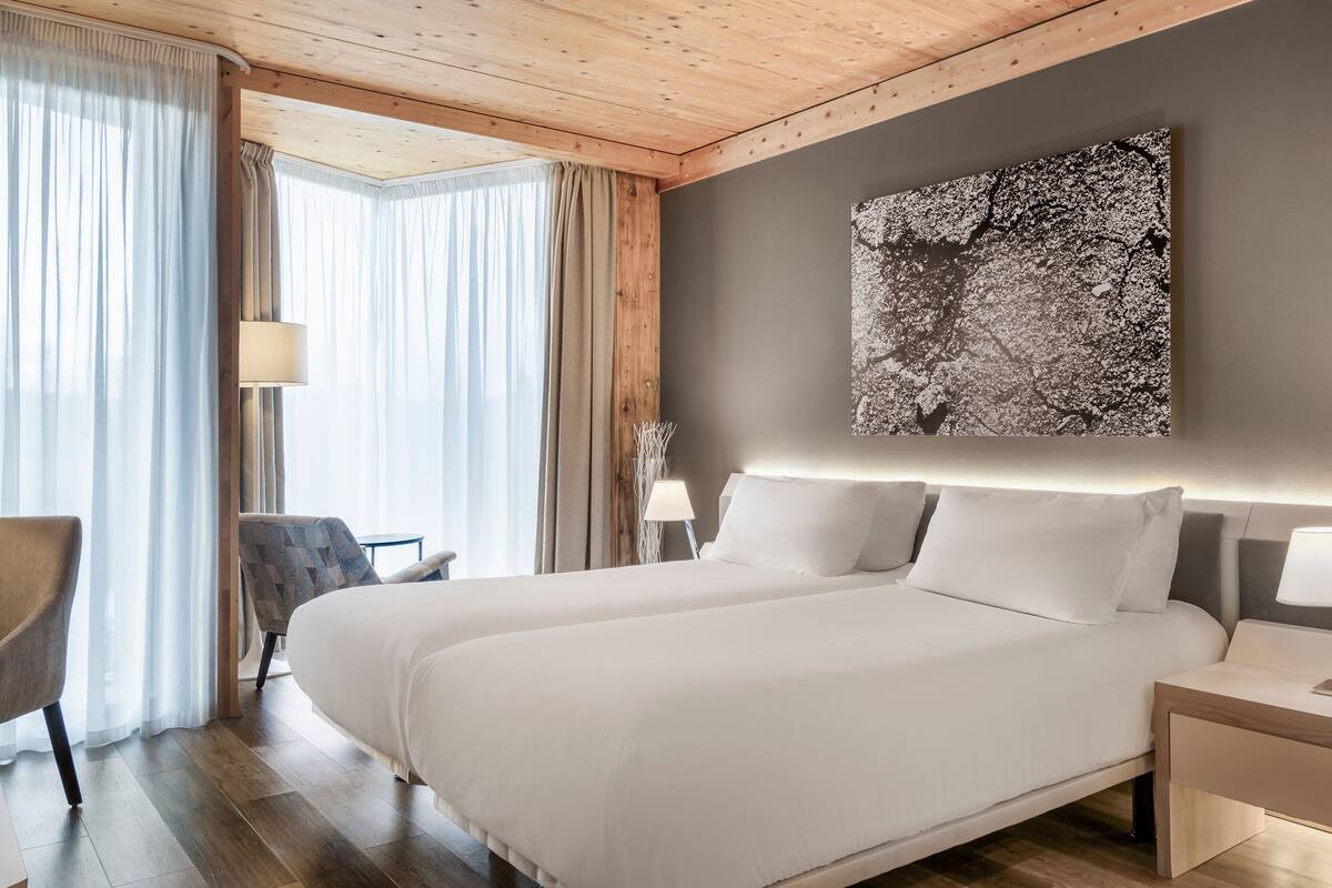 A modern hotel room with a large bed, two bedside tables, a chair, and a large window with curtains.