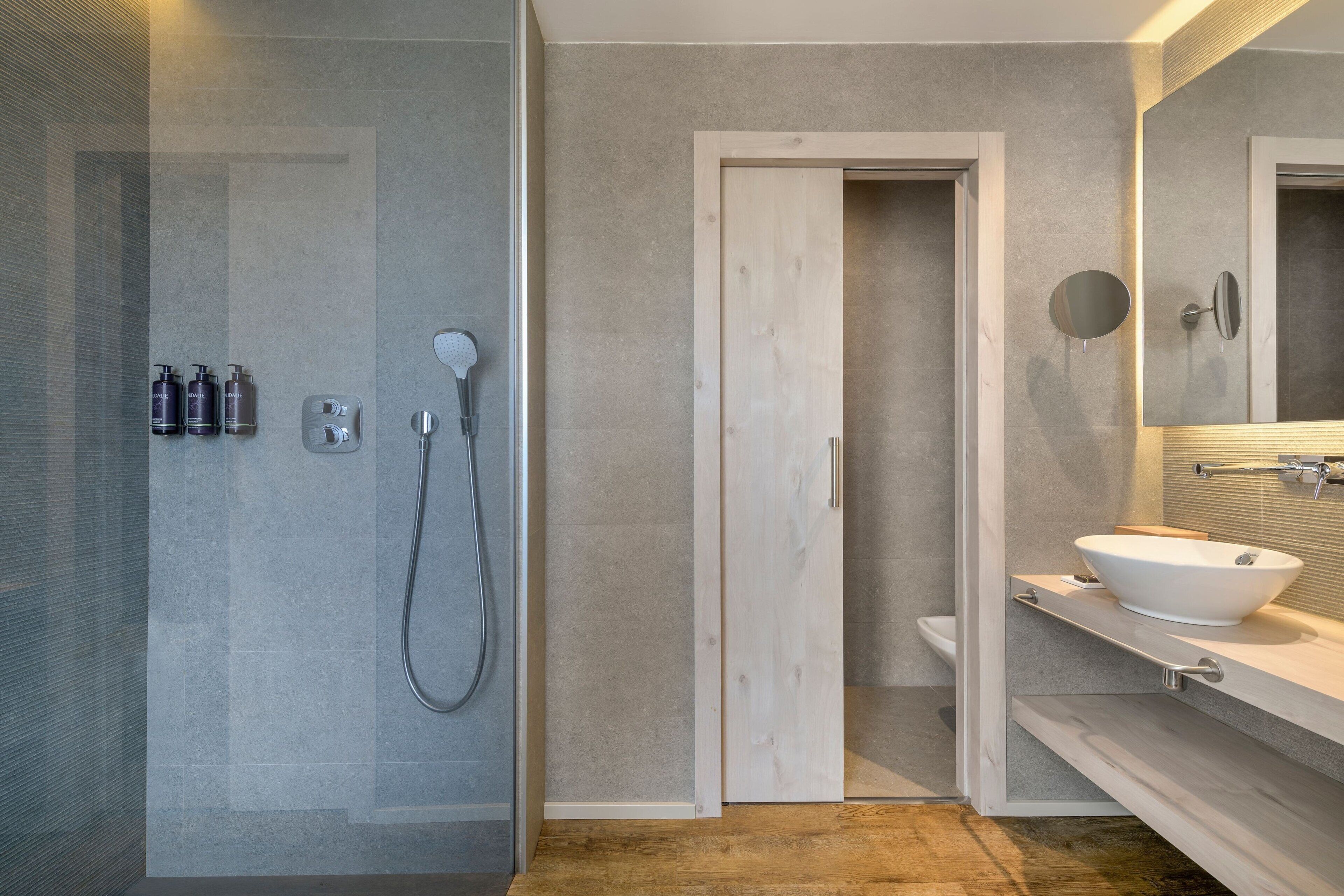 Un baño moderno con ducha de vidrio, puerta de madera, lavabo blanco y espejo con marco redondo.