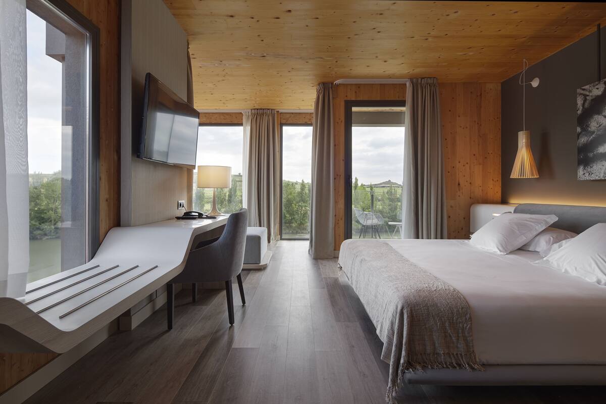 A modern hotel room with a wooden ceiling, a large bed, a desk with a chair, a TV, and a view of the outdoors.