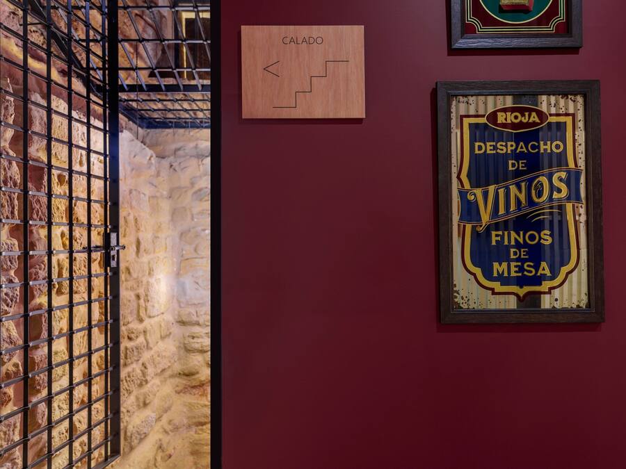 A stone wall with a metal gate, a wooden sign labeled "CALADO," and a framed sign with "RIOJA DESPACHO DE VINOS FINOS DE MESA."